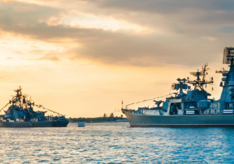 Naval ship cutting through ocean waves, with radar equipment visible against a blue sky.